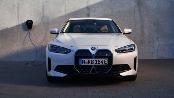 Näkymä keulasta ja BMW-kotilatauslaitteesta BMW i4 eDrive40 G26 2021 BMW i4 eDrive40 G26 2021 Mineral White näkymä keulasta talon seinän edessä jossa BMW-kotilatauslaite