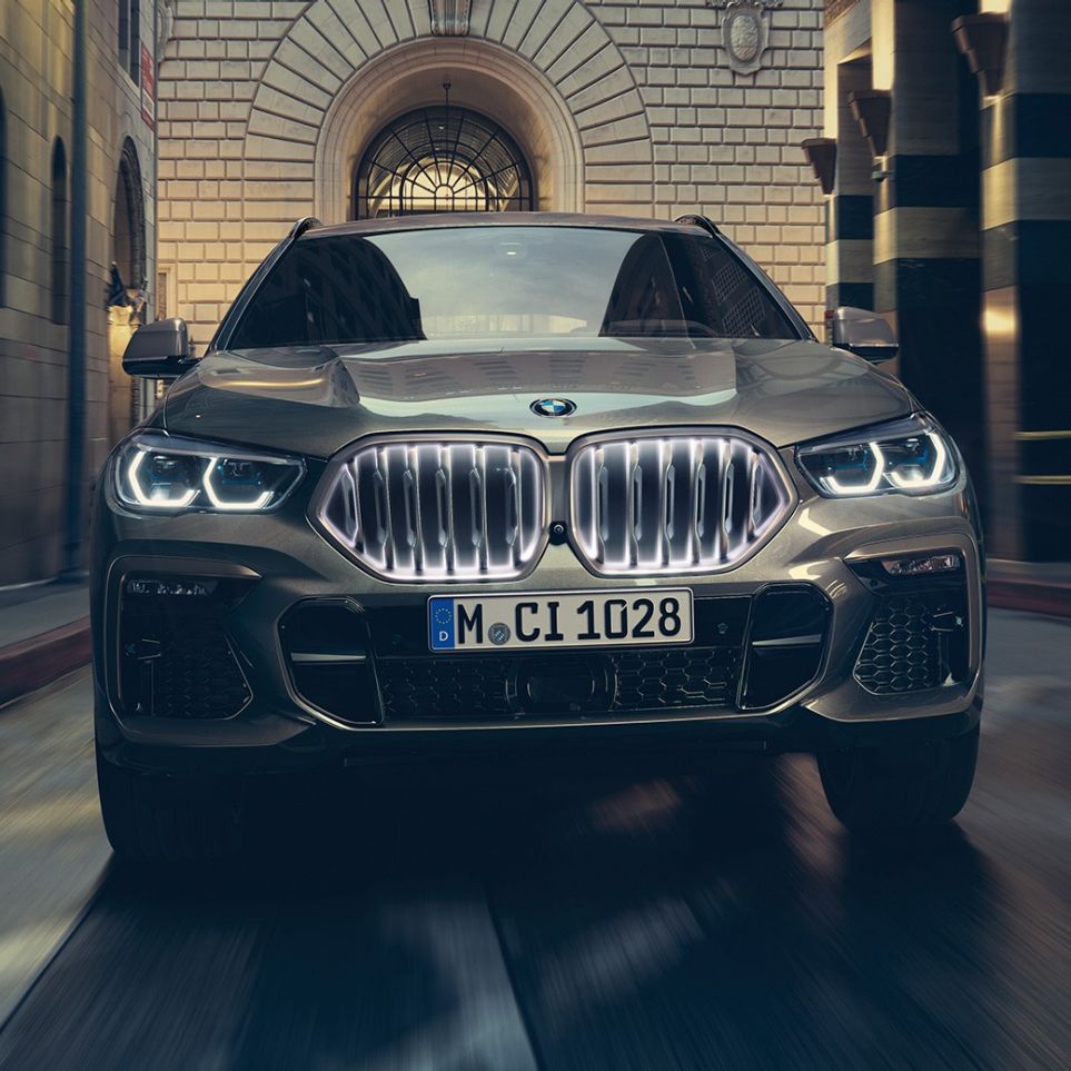 BMW X6, jossa BMW-jäähdyttimensäleikkö Iconic Glow. BMW X6, näkymä edestä kaupunkinäkymän edustalla.