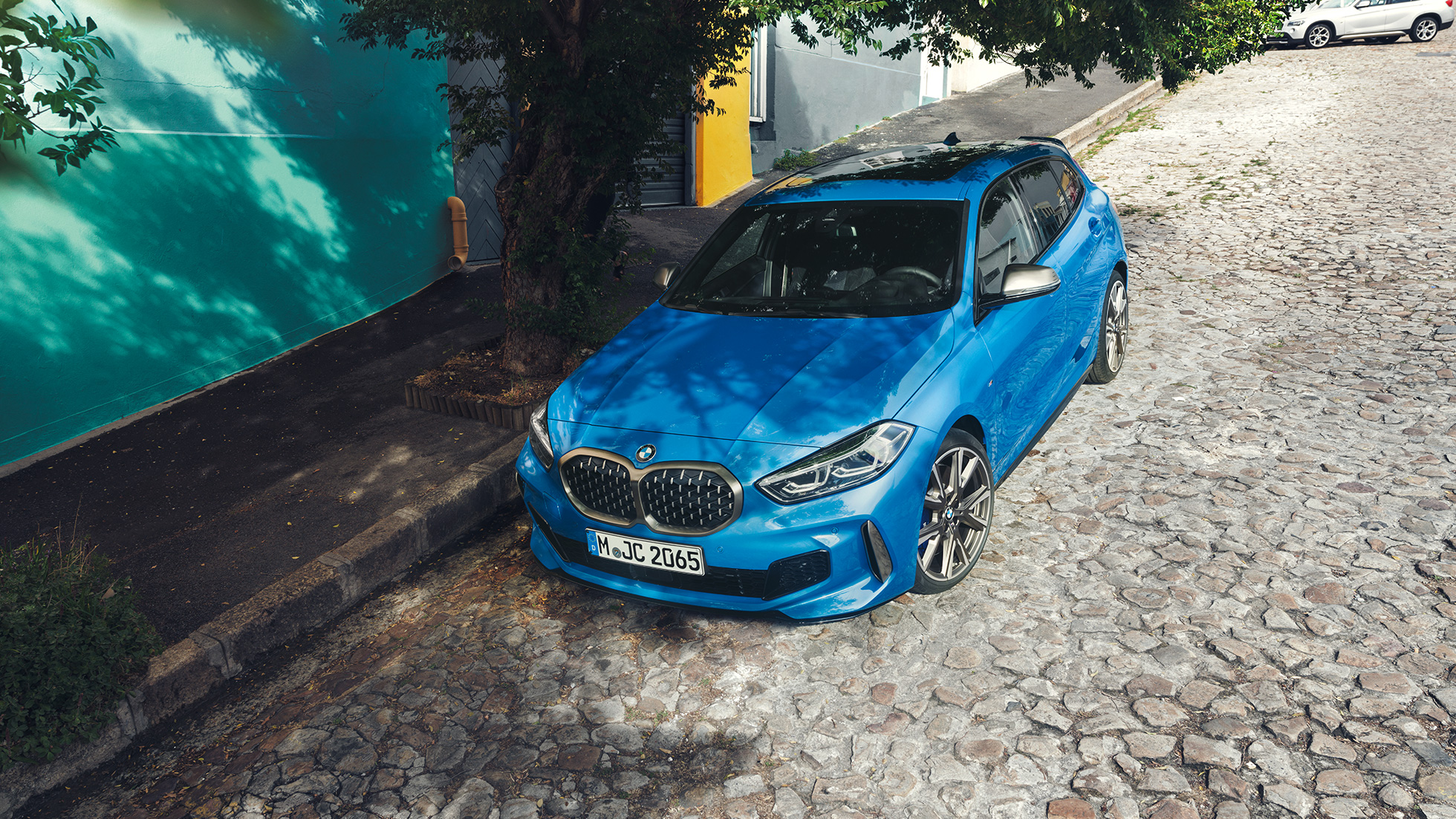 BMW 1-sarjan auto kadunvarteen pysäköitynä. BMW 1-sarjan auto, Misano Blue metalliväri, 3/4-näkymä etuosasta lintuperspektiivistä.
