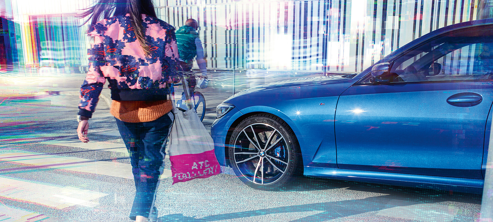BMW 3-sarjan Sedan, kolmeneljäsosa-sivukuva. Kolmeneljäsosa-sivukuva BMW 3-sarjan Sedanista asvaltilla suurkaupunkiympäristössä ohikulkijoineen.