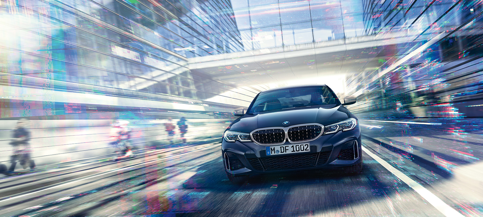BMW 3-sarjan Sedan, kuva BMW M340i xDrive -mallista edestäpäin. Lähikuva liikkuvasta BMW M340i xDrive Sedan -mallista edestäpäin suurkaupungin lasisten rakennusten välissä.
