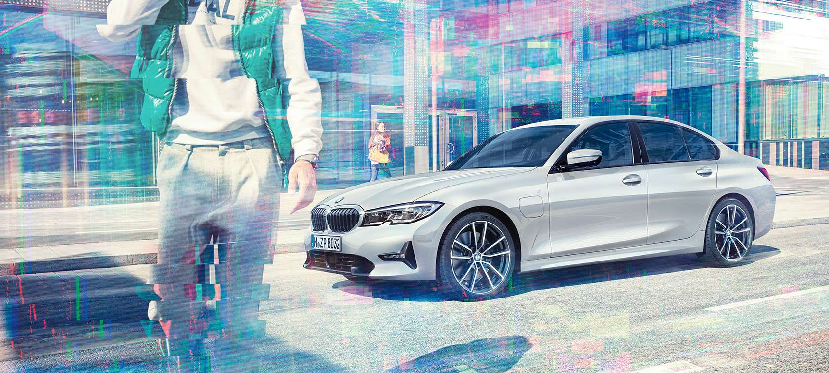 BMW 3-sarjan Sedan, sivukuva mallista BMW 330e. Kolmeneljäsosa-sivukuva paikallaan olevasta BMW 330e Sedanista suurkaupunki-lasirakennuksen edessä ohikulkijoineen.
