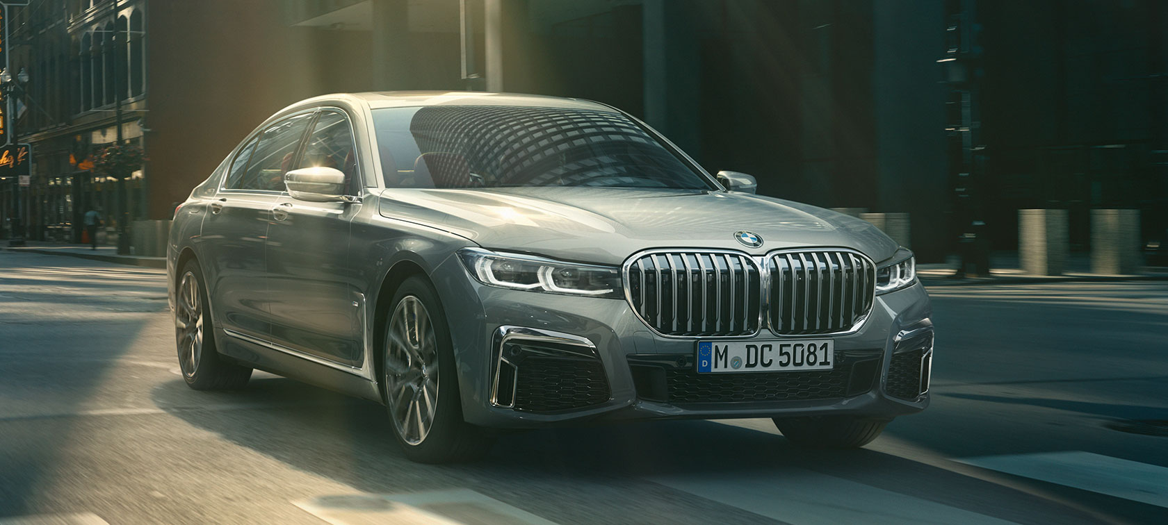 Harmaa BMW 7-sarjan Sedan ajaa, etuosan kolme neljäsosaa Harmaa BMW 7-sarjan Sedan ajaa kaupunkimaisemassa, etuosasta näkyy kolme neljäsosaa