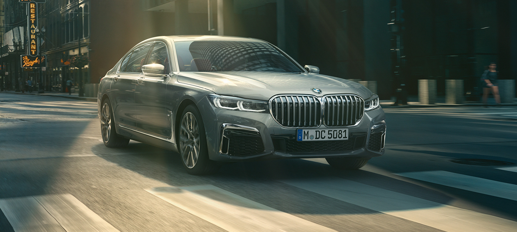 Harmaa BMW 7-sarjan Sedan ajaa, etuosan kolme neljäsosaa Harmaa BMW 7-sarjan Sedan ajaa kaupunkimaisemassa, etuosasta näkyy kolme neljäsosaa