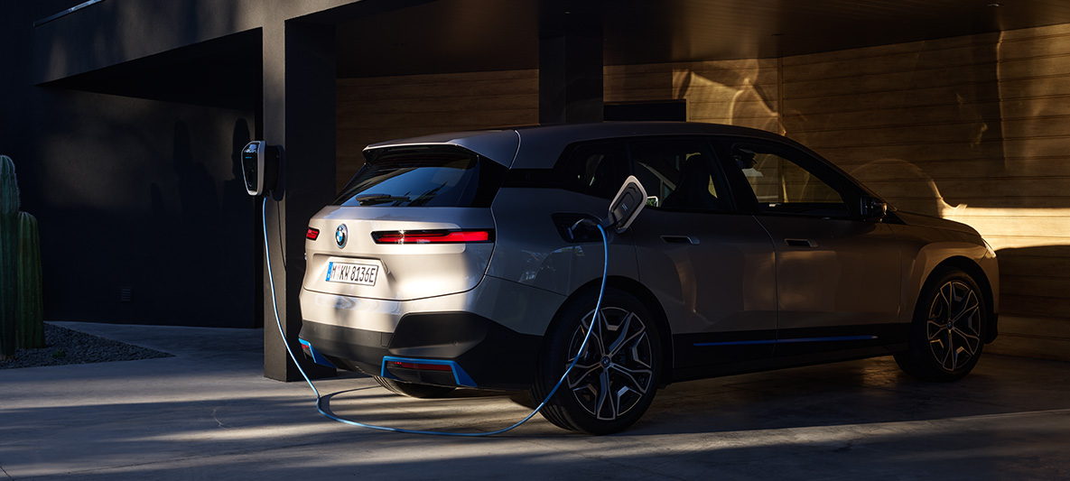 BMW iX, kolmen neljäsosan näkymä perästä, kun autoa ladataan BMW Wallbox -latauslaitteella BMW iX Cashmere Silver i20, kolmen neljäsosan näkymä perästä, kun autoa ladataan BMW Wallbox -latauslaitteella