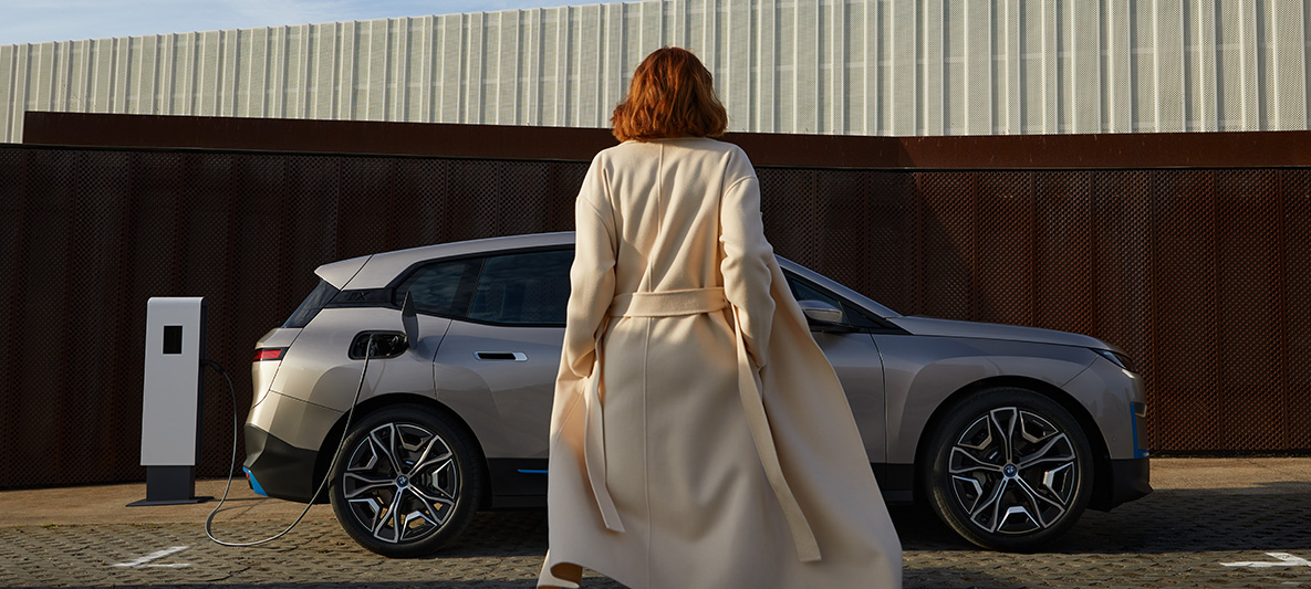 BMW iX, sivunäkymä pysähtyneenä olevasta autosta BMW iX Cashmere Silver i20, sivunäkymä pysähtyneenä olevasta autosta, jonka edessä seisoo malli