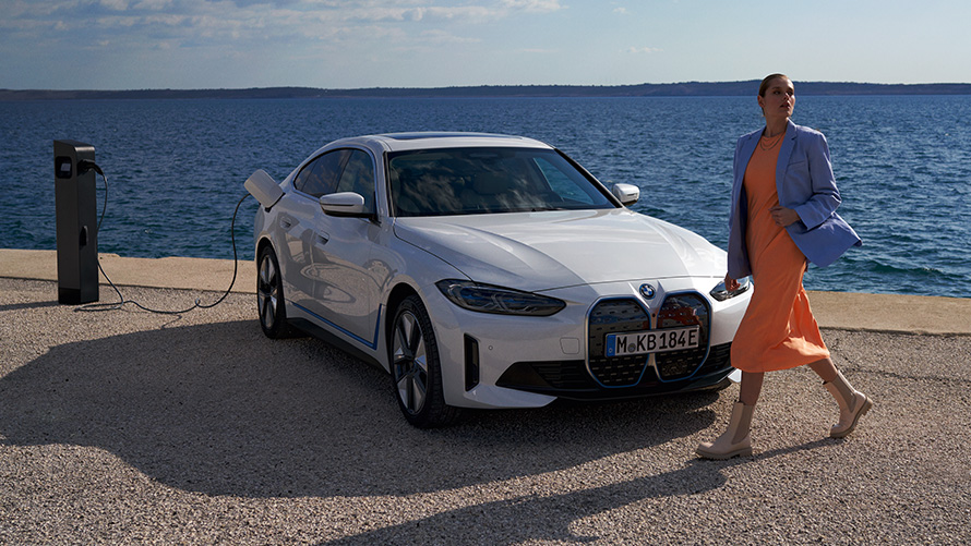 Kolmen neljäsosan näkymä edestä i4 eDrive40 G26 2021 latausasemalla Nainen BMW i4 eDrive40 G26 2021:n edessä Mineral White kolmen neljäsosan näkymä edestä latauspisteellä järven edessä