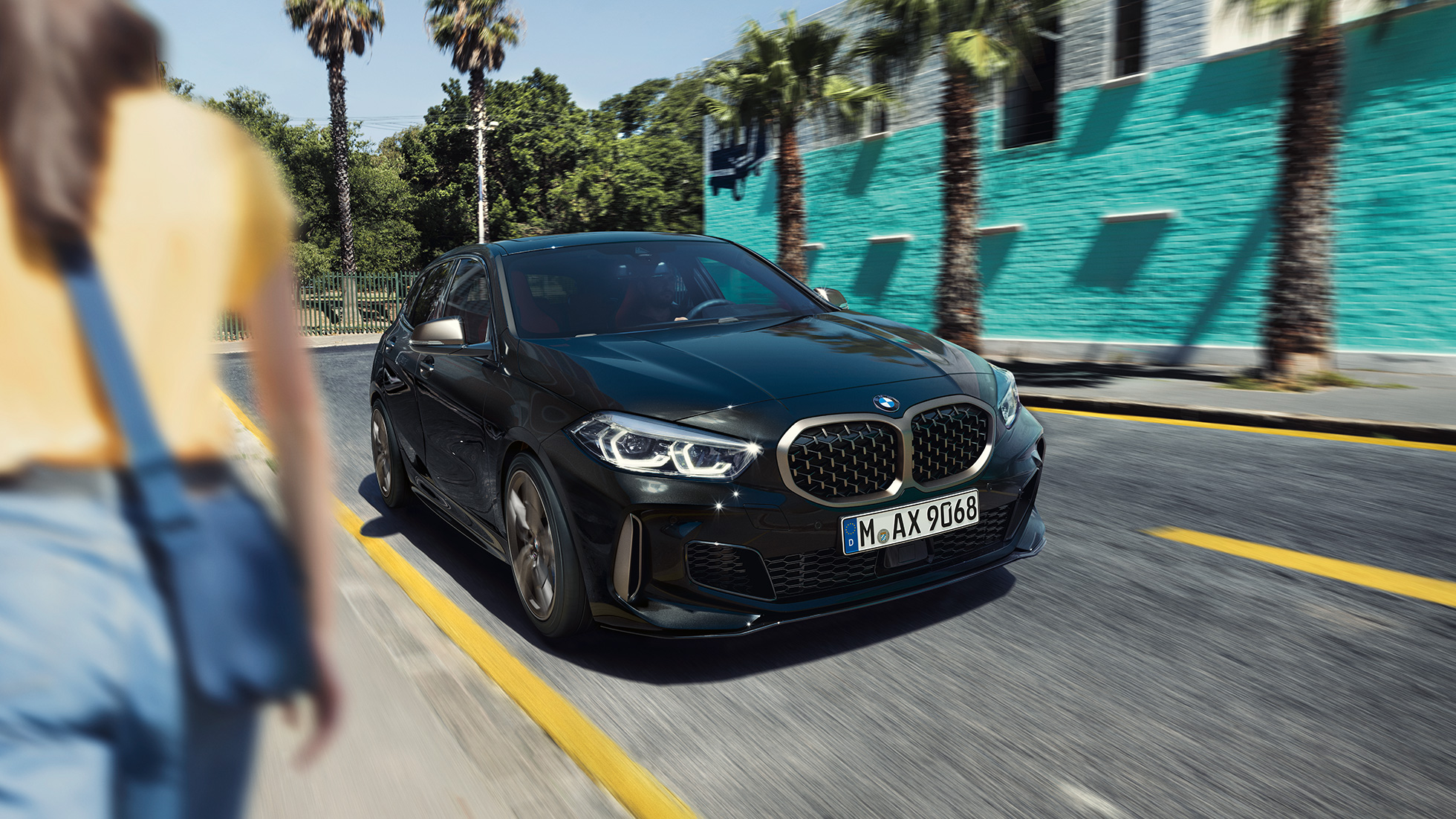 BMW M135i xDrive, 3/4 näkymä keulasta. BMW M135i xDrive, metalliväri Sapphire Black, 3/4 etunäkymä ajavasta autosta nainen etualalla.