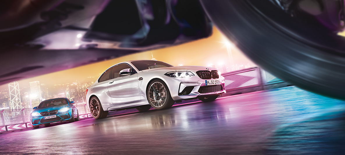 BMW M2 Competition heijastavalla asfaltilla Kaksi BMW M2 Competition -autoa kilparadalla taustallaan öinen suurkaupunkimaisema maantasosta kuvattuna.