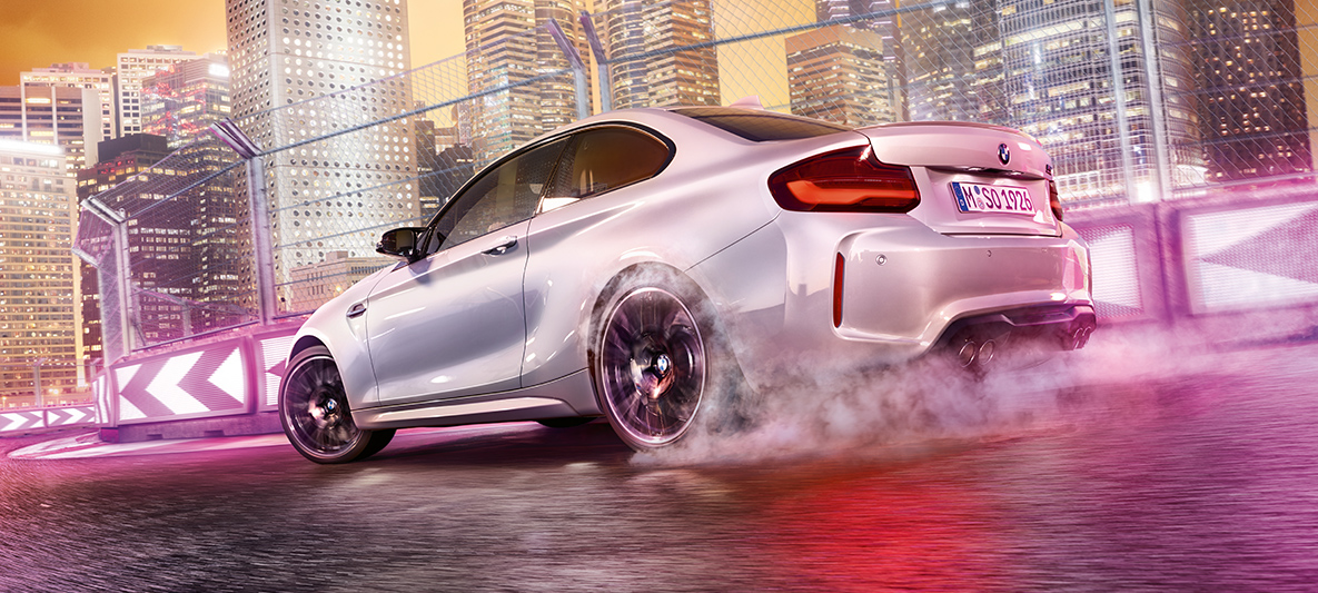 BMW M2 Competition kiihdyttää Valkoinen BMW M2 Competition kiitää kilparadalla taustallaan öinen maisema.