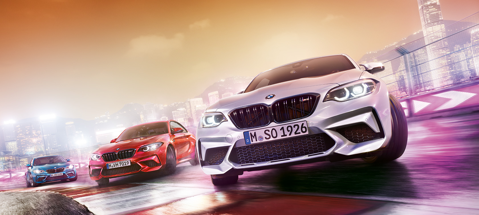 BMW M2 Competition toiminnassa Kolme BMW M2 Competition -autoa kiitää kilparadalla taustallaan öinen suurkaupunkimaisema.