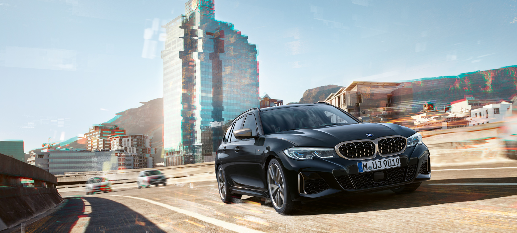 Kolme neljäsosan näkymä edestä auton ajaessa kaupunkinäkymän edessä BMW M340i xDrive Touring, kolme neljäsosan näkymä edestä auton ajaessa kaupunkinäkymän edessä
