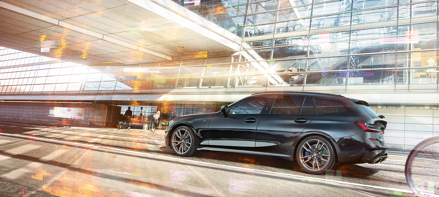 BMW M340i xDrive Touring, sivukuva liikkeellä olevasta autosta BMW M340i xDrive Touring, sivukuva urbaanin lasirakennuksen edessä ajavasta autosta