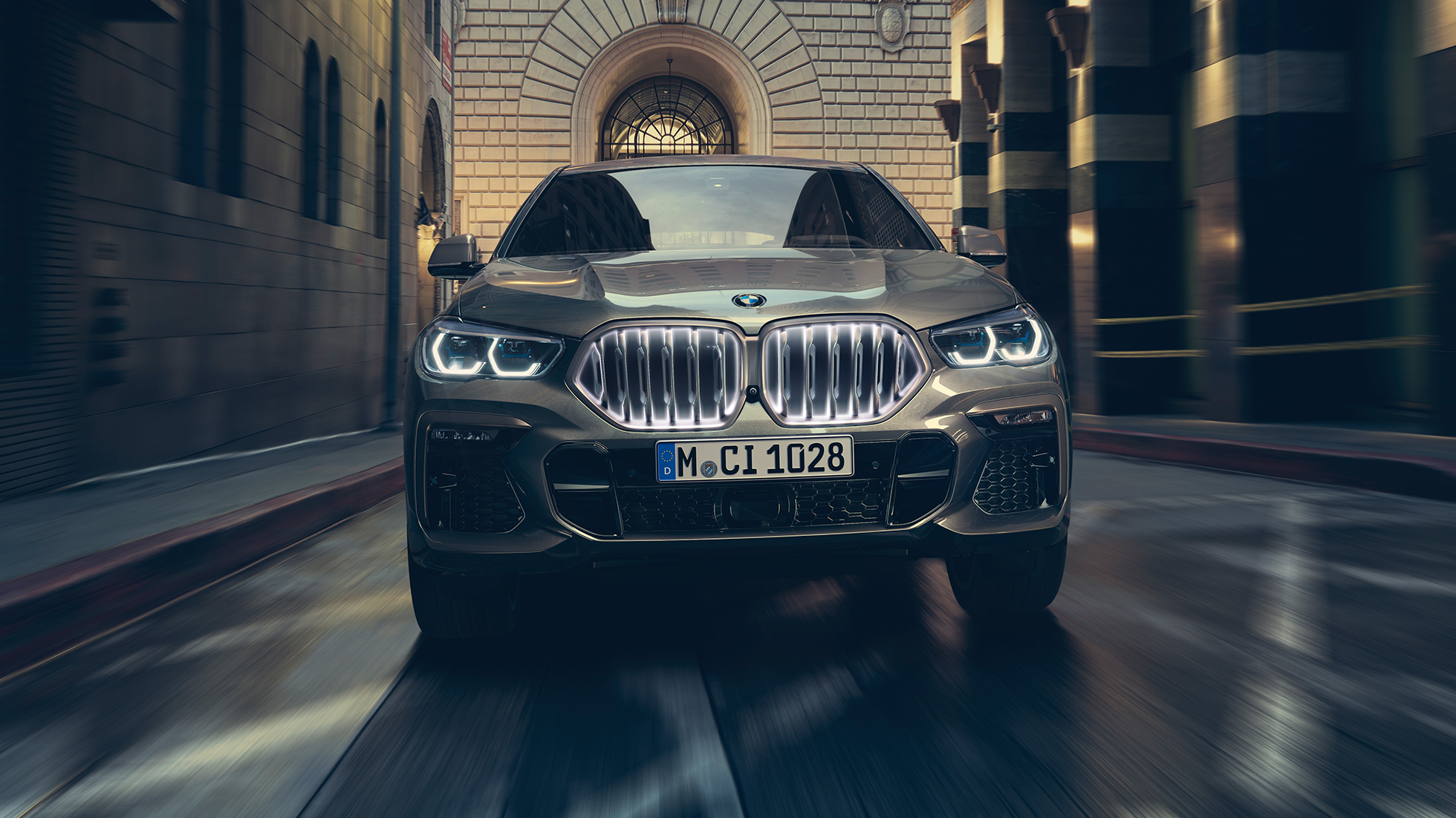 BMW X6 M50i, jossa BMW-jäähdyttimensäleikkö Iconic Glow. BMW X6 M50i, näkymä edestä auton ollessa kaupunkinäkymän edustalla.