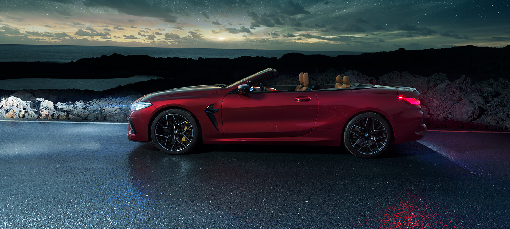 BMW M8 Competition Cabrio, sivunäkymä. BMW M8 Competition Cabrio Motegi Red -metallivärisenä ja katto avattuna, sivunäkymä paikallaan olevasta autosta rantatiellä tähtitaivaan alla.