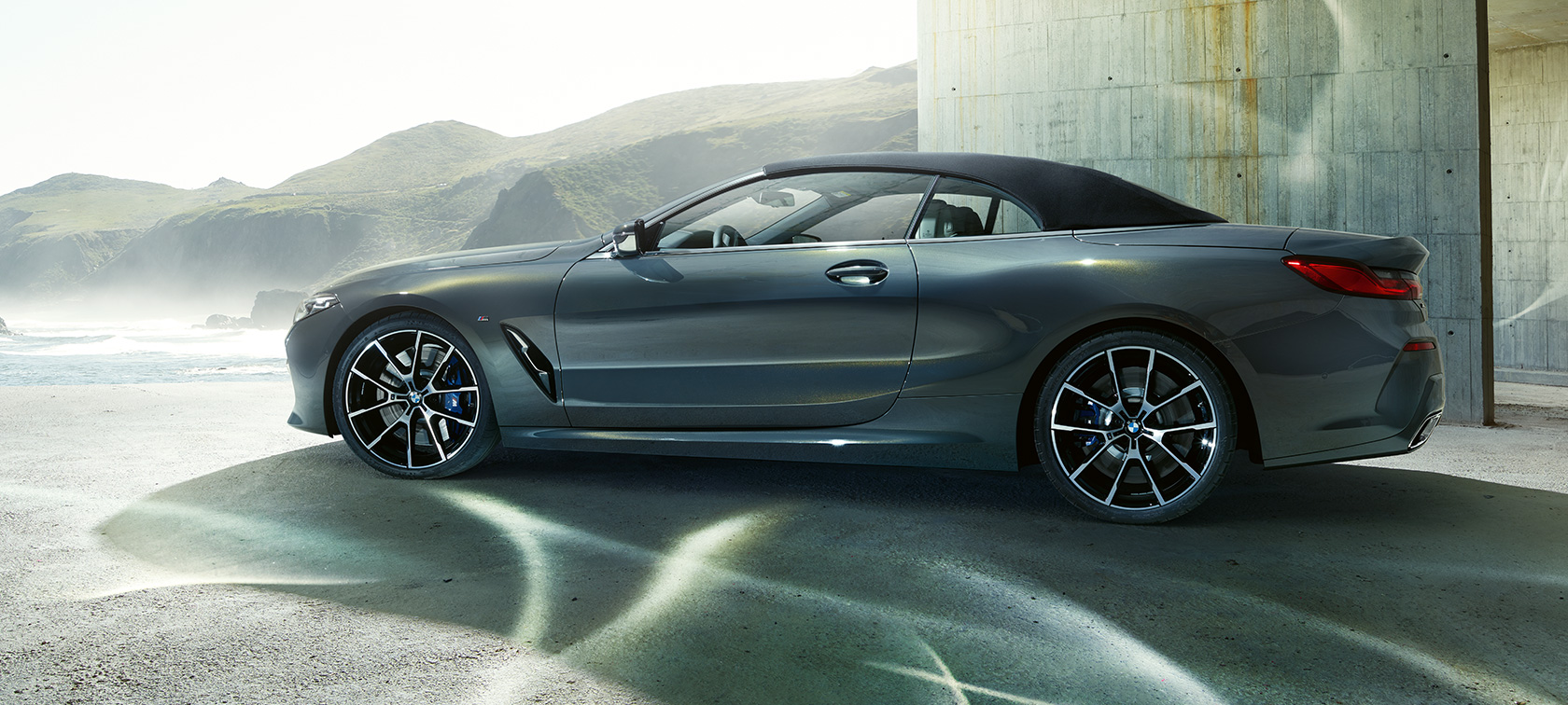 BMW M850i xDrive Cabrio, sivunäkymä. BMW M850i xDrive Cabrio Dravite Grey -metallivärisenä ja kangaskatto suljettuna, sivunäkymä kukkuloiden edustalla.