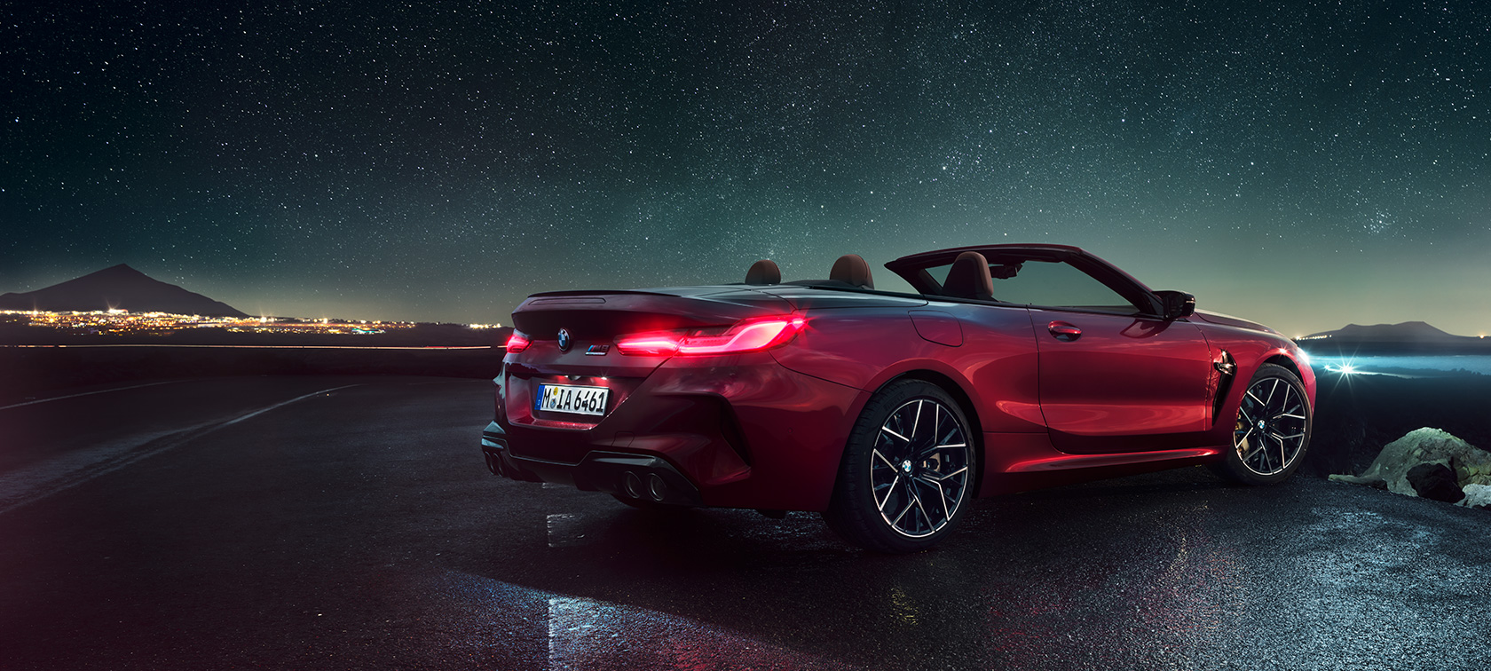 BMW M8 Competition Cabrio, 3/4-näkymä takaa. BMW M8 Competition Cabrio Motegi Red -metallivärisenä ja katto avattuna, 3/4-näkymä takaa paikallaan olevasta autosta tähtitaivaan alla.