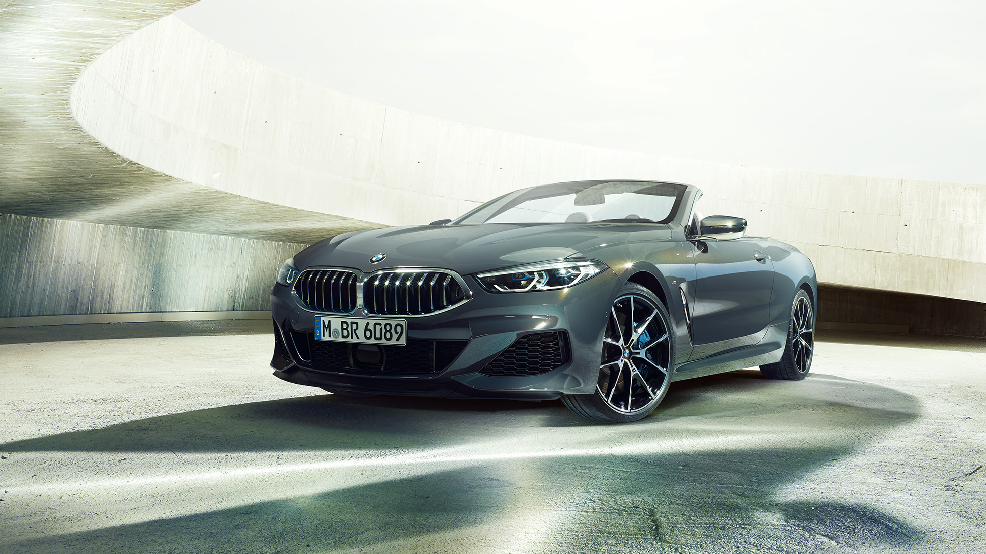 BMW M850i xDrive Cabrio, 3/4-näkymä edestä. BMW M850i xDrive Cabrio Dravite Grey -metallivärisenä ja katto avattuna, 3/4-näkymä edestä auton ollessa pysähtyneenä betonirakennuksessa.