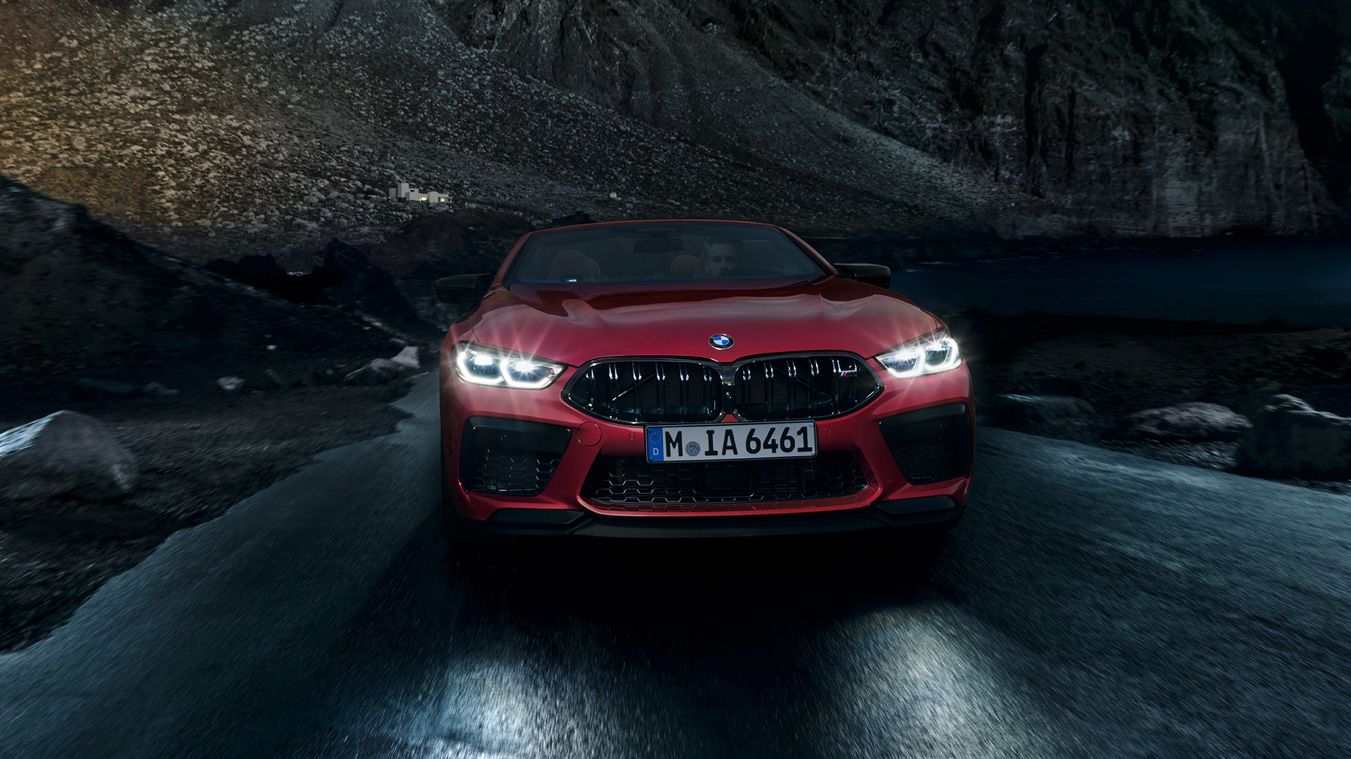BMW M8 Competition Cabrio, näkymä edestä. BMW M8 Competition Cabrio Motegi Red -metallivärisenä ja ajovalojen palaessa, näkymä edestä auton ollessa liikkeellä yöllä.