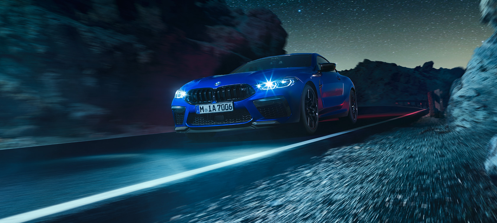 BMW M8 Competition Coupé, 3/4-näkymä edestä. BMW M8 Competition Coupé metallivärissä Marina Bay Blue, 3/4-näkymä edestä auton ollessa liikkeellä yöllä.