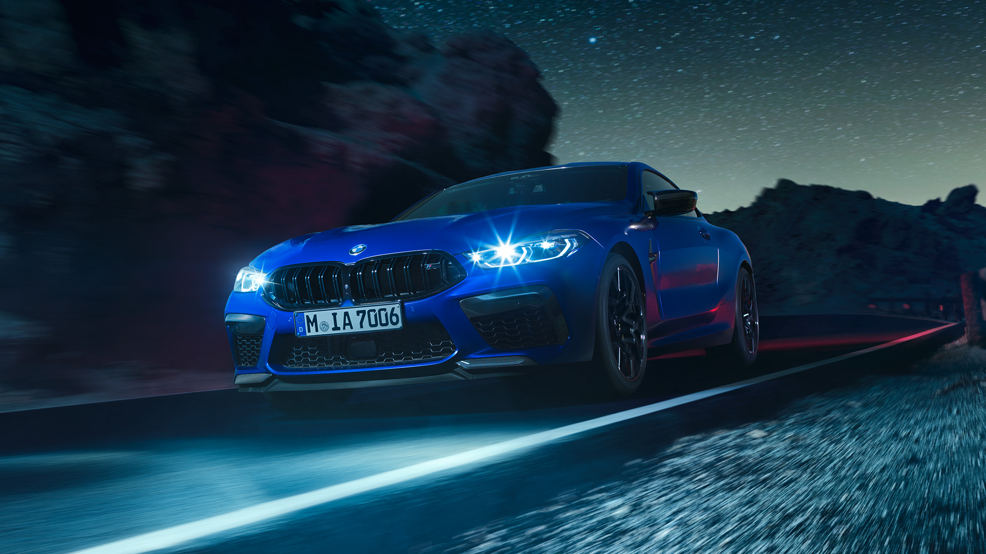 Liikkeellä oleva BMW M8 Competition Coupé, 3/4-näkymä edestä. BMW M8 Competition Coupé metallivärissä Marina Bay Blue ja LED-ajovalojen palaessa, 3/4-näkymä yöllä liikkeellä olevan auton etuosasta.