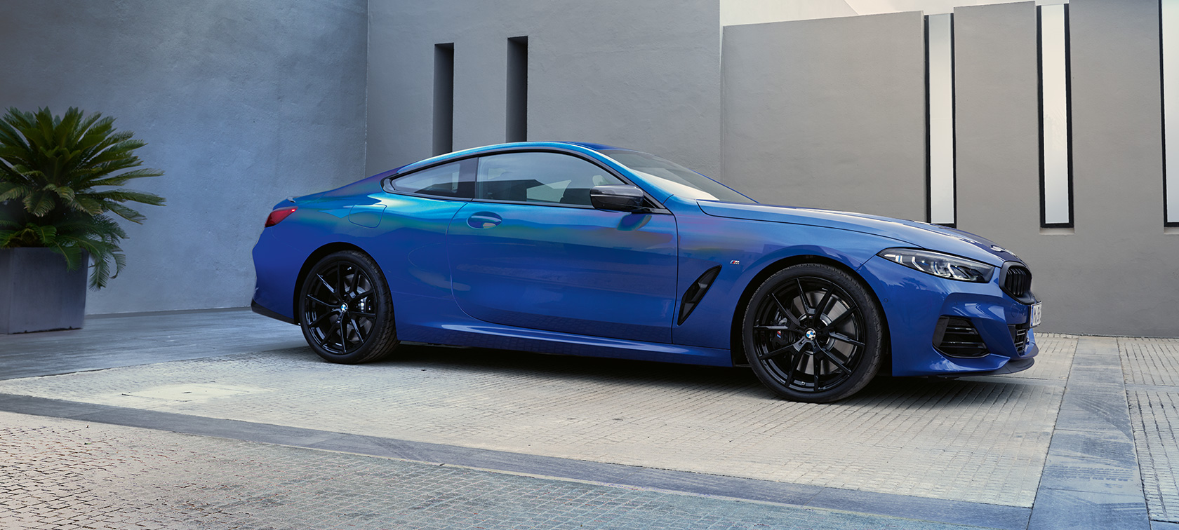 Sivunäkymä BMW M850i xDrive Coupé G15 LCI Facelift 2022 BMW M850i xDrive Coupé G15 LCI Facelift 2022 M Portimao Blau -metalliväri sivunäkymänä auton ollessa pysähdyksissä rakennuksen edessä