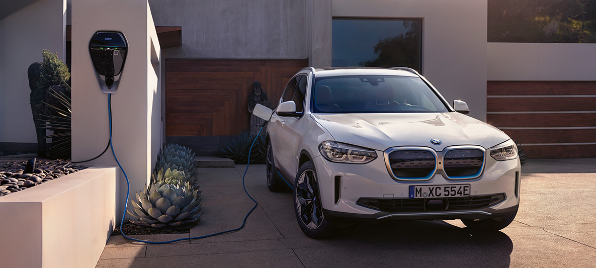 BMW iX3 G08, kolmen neljäsosan näkymä keulasta BMW Wallboxin luona BMW iX3 G08 2020 metalliväri Mineral White, kolmen neljäsosan näkymä keulasta BMW Wallbox -latauspisteen luona talon edessä
