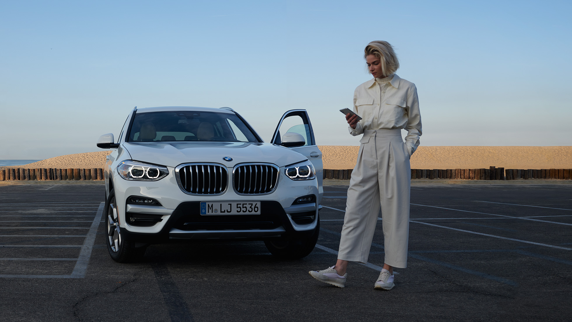 Nainen käyttää My BMW -sovellusta älypuhelimella Nainen käyttää BMW ConnectedDriven My BMW -sovellusta äypuhelimella valkoisen BMW X5 -auton edessä