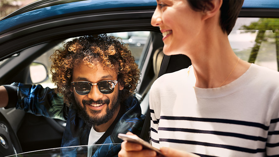 BMW Connected BMW:n sisällä oleva mies ja BMW:n ulkopuolella oleva nainen käyttävät BMW Connected -palveluja älypuhelimella