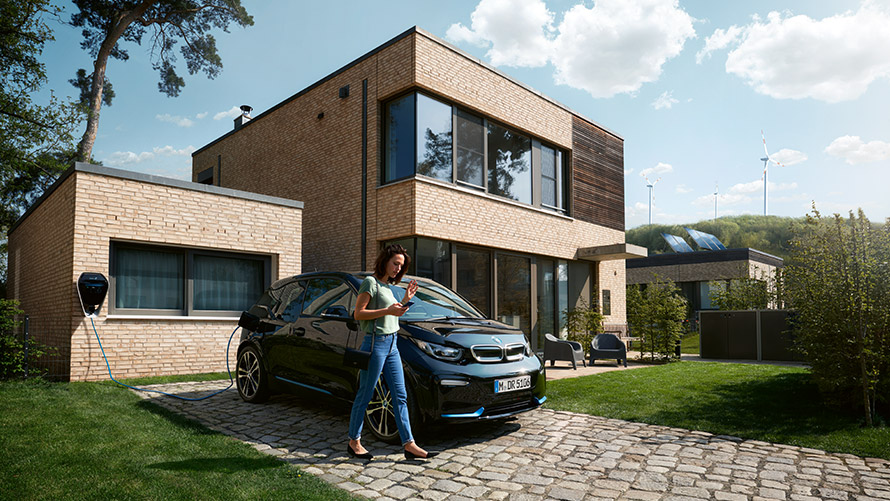 Digital Charging Service (ei saatavilla Suomessa) Nainen kävelee ulkona BMW i3 -autonsa edessä ja käyttää Digital Charging Service -toimintoa älypuhelimellaan