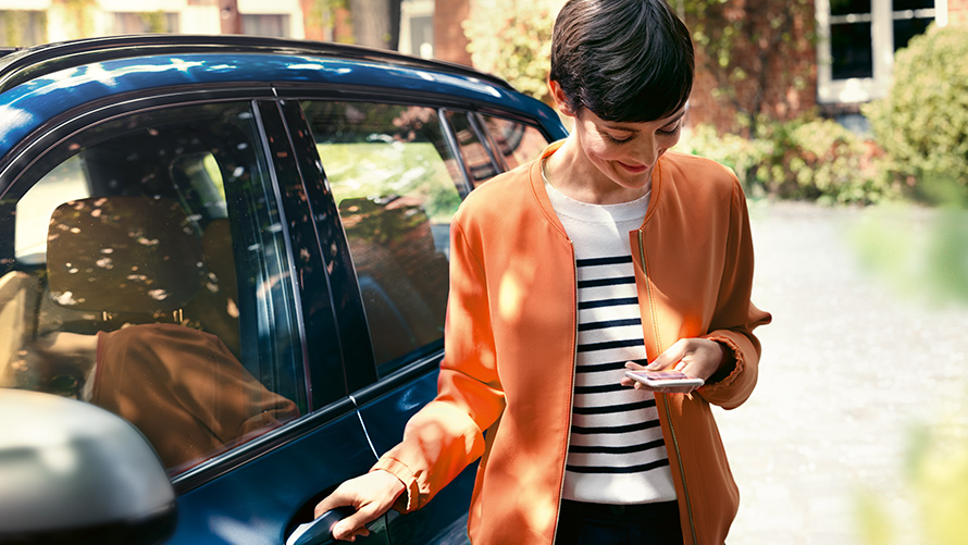 BMW Teleservices Nainen seisoo BMWnsä oven luona älypuhelimen kanssa