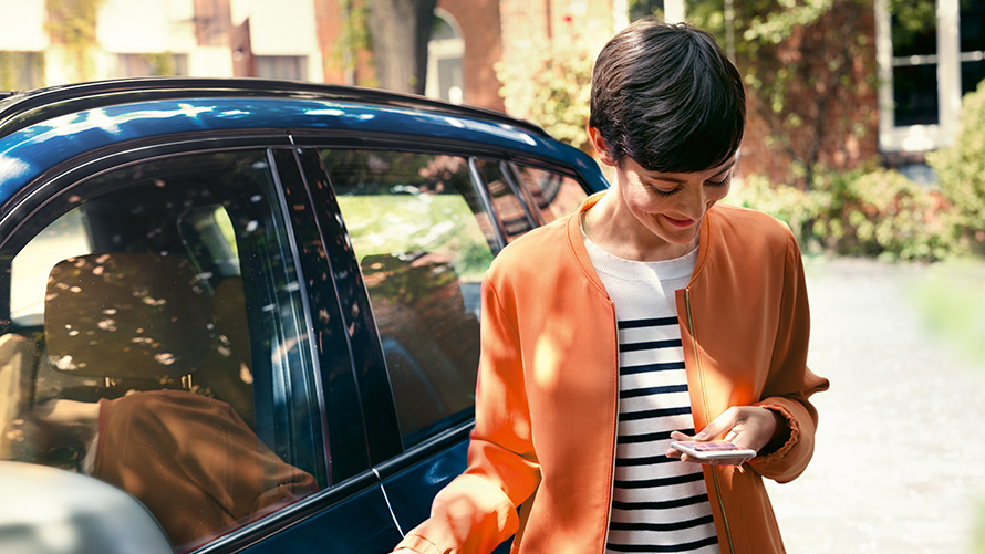 BMW-kesätarjoukset, BMW Teleservices BMW-kesätarjoukset, BMW Teleservices, nainen seisoo BMW-auton edessä ja käyttää älypuhelinta